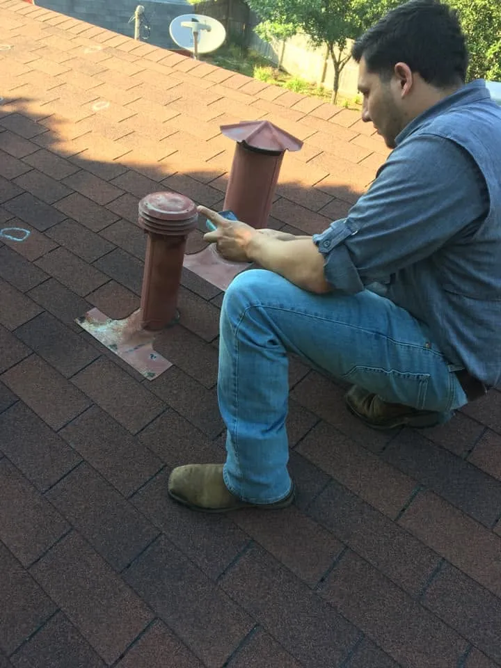 Braulio Lozano kneeling on a shingle roof, taking notes on his phone beside two roof vents