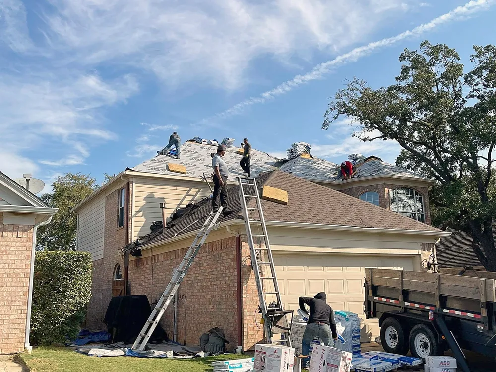 Roofers on ladders and on the roof of a two-story home, bundles of shingles and a trailer in the drive