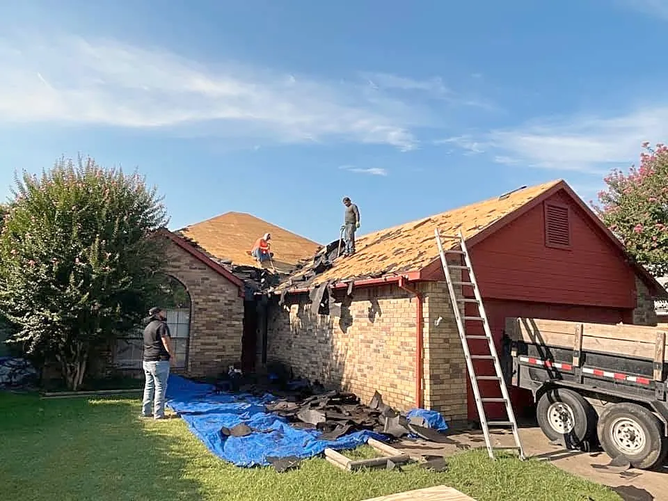 A crew tearing off an old roof down to the deck, with a dump trailer in the drive